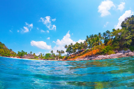 Bright sunlight illuminates the vibrant turquoise waters as gentle waves lap against a sandy beach surrounded by towering palm trees and lush greenery.の写真素材