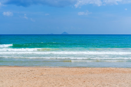 Waves gently crash on a sandy shore while two small islands are visible on the horizon. The sky is mostly clear with a few clouds, creating a tranquil beach atmosphere.の写真素材