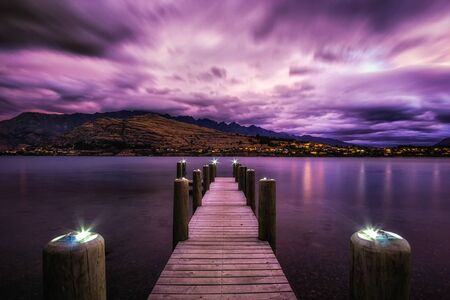 Sunset over lake Wakatipu with kelvin heights in the distance. Sunlight covered in stormy clouds. Taken during summer. Small dock in the foreground.の写真素材
