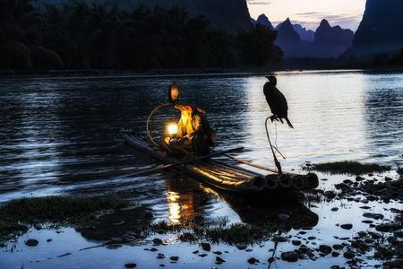 Li river cormant fisherman resting by the famous li river with the 20 yuan bank note scenery on the background. Taken near Xingping, which is nearby yangshuo in guilin. The fisherman has a small lantern on the bamboo raft.の写真素材