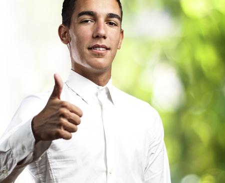 young man with thumb up wearing white shirt against a green parkの写真素材