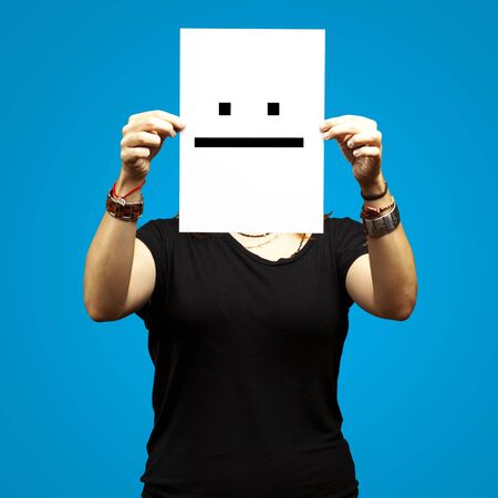 woman holding paper with poker face emoticon against a blue backgroundの写真素材