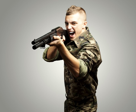 portrait of a young soldier aiming with shotgun over grey background