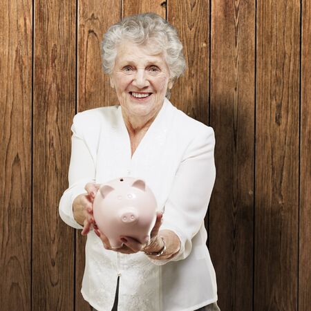 portrait of senior woman showing a piggy bank against a wooden wallの写真素材