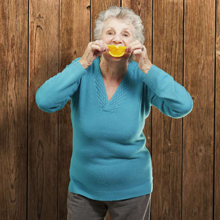 portrait of senior woman holding a orange slice in front of her mouth against a wooden wallの写真素材