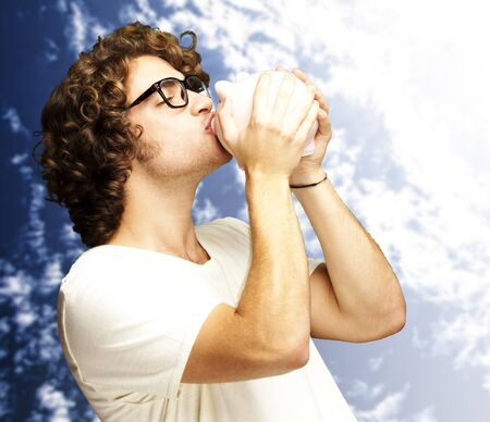 portrait of young man kissing a piggy bank against a cloudy sky backgroundの写真素材