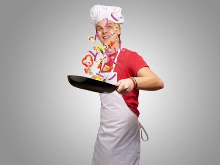 portrait of young cook man cooking vegetables on pan over grey backgroundの写真素材