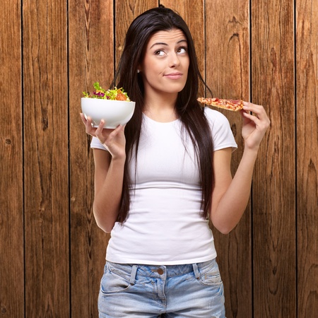 portrait of young woman choosing pizza or salad against a wooden wallの写真素材