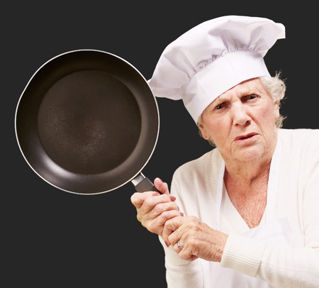 Cook senior woman angry trying to hit with pan over black backgroundの写真素材