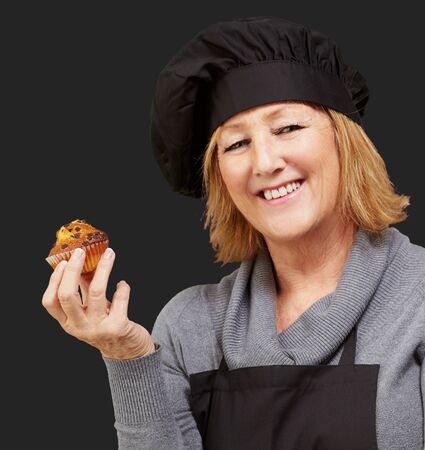 portrait of middle aged cook woman holding a delicious homemade muffin over black backgroundの写真素材
