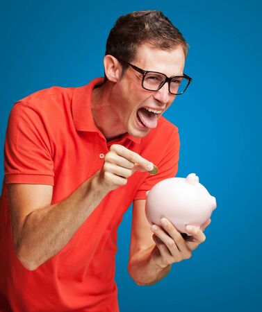 Portrait Of  Man With Piggy Bank Inserting A Coin On Blue Backgroundの写真素材