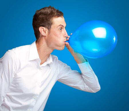 Portrait Of Young Man Blowing A Balloon Isolated On Blue Background