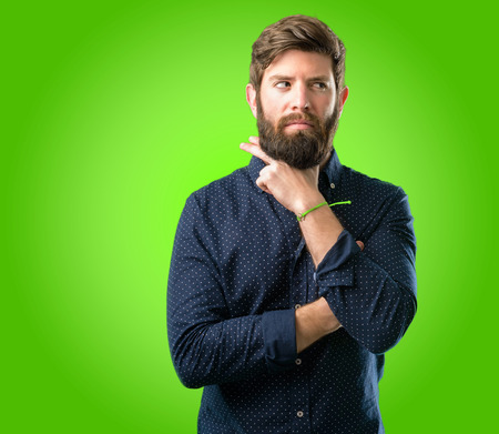 Young hipster man with big beard thinking thoughtful with smart face, expressing question and doubt. Imagine the solution over green backgroundの写真素材