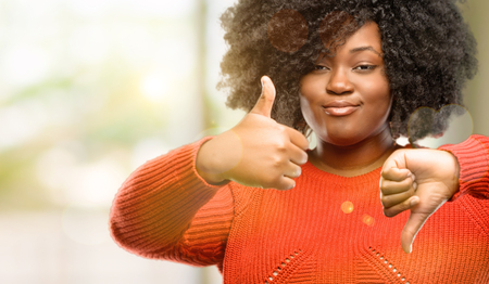 Beautiful african woman confused with thumbs up and down, trying to take a decision expressing doubt and frustration, outdoorの写真素材