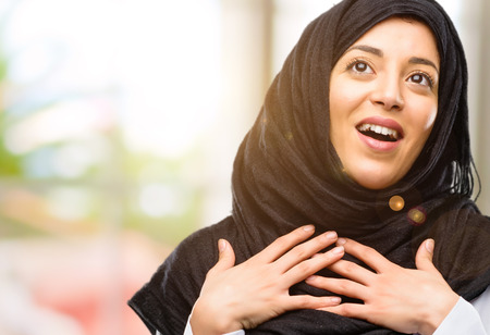 Young arab woman wearing hijab having charming smile holding hands on heart wanting to show love and sympathyの写真素材