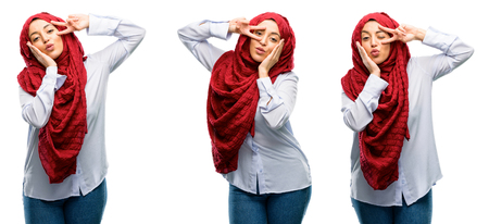 Arab woman wearing hijab looking at camera through her fingers in victory gesture winking the eye and blowing a kiss isolated over white backgroundの写真素材