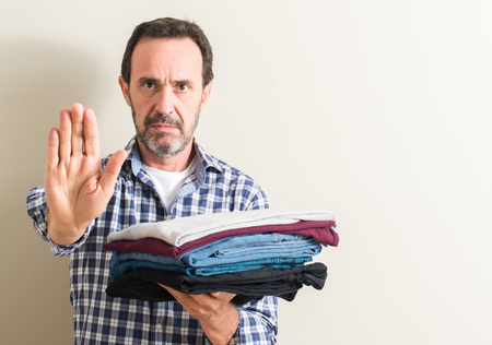 Senior man holding folded laundry clothes with open hand doing stop sign with serious and confident expression, defense gestureの写真素材