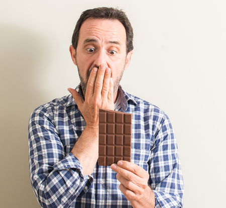 Senior man eating chocolate bar cover mouth with hand shocked with shame for mistake, expression of fear, scared in silence, secret conceptの写真素材