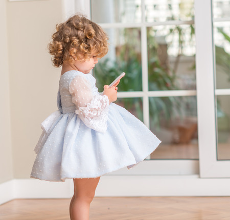 Beautiful blonde child in a dress standing playing with smartphone at home.の写真素材