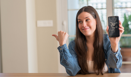 Brunette young woman holding broken smartphone pointing with hand and finger up with happy face smilingの写真素材