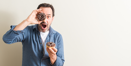 Senior man eating chocolate donut scared in shock with a surprise face, afraid and excited with fear expressionの写真素材
