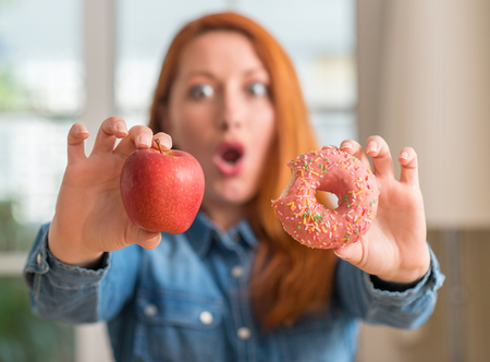 Redhead woman chooses between apple and donut scared in shock with a surprise face, afraid and excited with fear expressionの写真素材