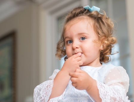 Beautiful blond child in a dress eating candy with hand on mouth at homeの写真素材