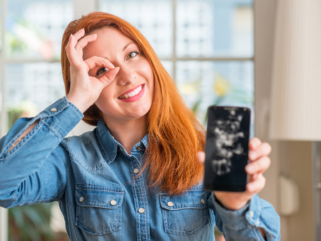 Redhead woman holding broken smartphone with happy face smiling doing ok sign with hand on eye looking through fingersの写真素材