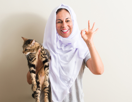 Arabian woman wearing white hijab holding a cat doing ok sign with fingers, excellent symbolの写真素材