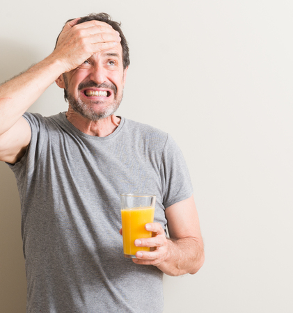 Senior man drinking orange juice in a glass stressed with hand on head, shocked with shame and surprise face, angry and frustrated. Fear and upset for mistake.の写真素材