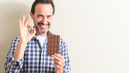 Senior man eating chocolate bar doing ok sign with fingers, excellent symbolの写真素材