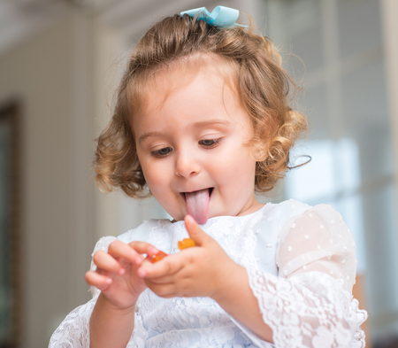 Beautiful blond child in a dress eating candy with tongue out at home.