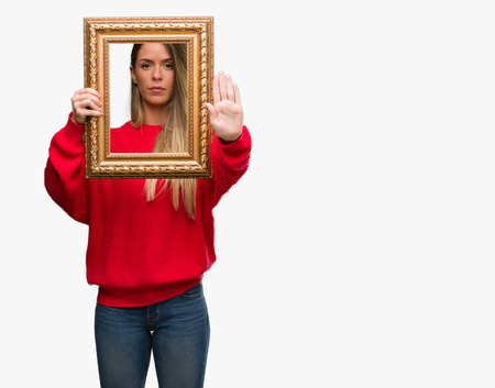 Beautiful young woman holding vintage frame with open hand doing stop sign with serious and confident expression, defense gestureの写真素材