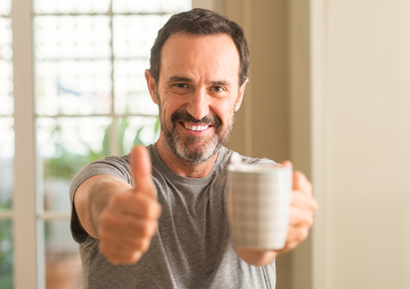 Middle age man drinking coffee in a cup happy with big smile doing ok sign, thumb up with fingers, excellent signの写真素材