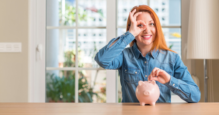 Redhead woman saves money in piggy bank at home with happy face smiling doing ok sign with hand on eye looking through fingersの写真素材