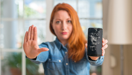 Redhead woman holding broken smartphone with open hand doing stop sign with serious and confident expression, defense gestureの写真素材