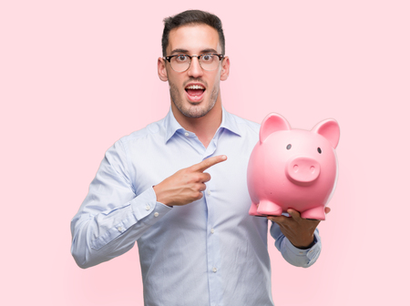 Handsome young man holding a piggy bank very happy pointing with hand and fingerの写真素材