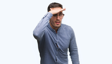 Handsome young elegant man wearing glasses very happy and smiling looking far away with hand over head. Searching concept.の写真素材