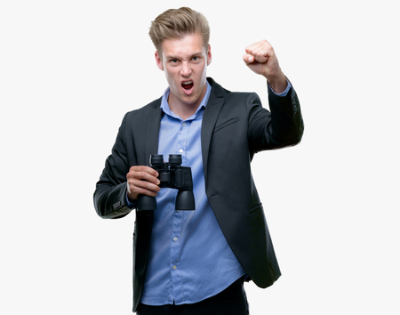 Young handsome blond man looking through binoculars annoyed and frustrated shouting with anger, crazy and yelling with raised hand, anger conceptの写真素材