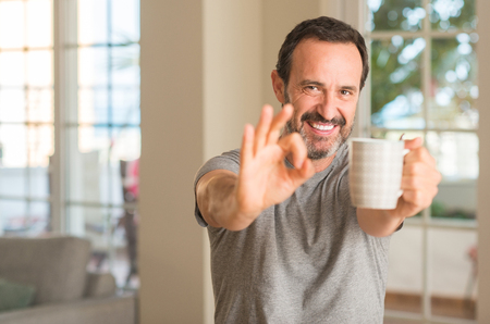 Middle age man drinking coffee in a cup doing ok sign with fingers, excellent symbolの写真素材