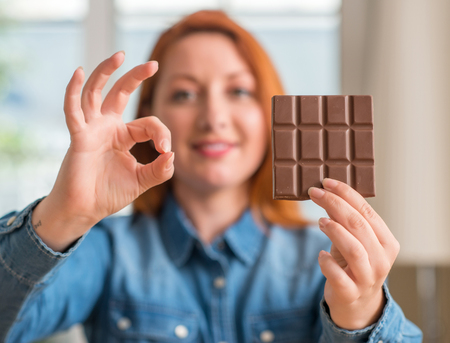 Redhead woman holding chocolate bar at home doing ok sign with fingers, excellent symbol