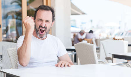 Handsome senior man sitting at terrace annoyed and frustrated shouting with anger, crazy and yelling with raised hand, anger conceptの写真素材
