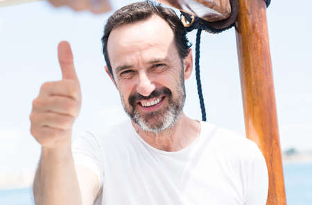 Handsome senior man traveling on sailboat happy with big smile doing ok sign, thumb up with fingers,...