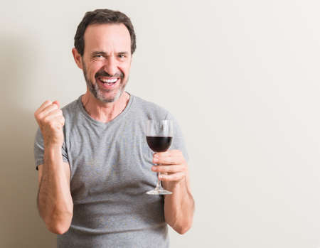 Senior man drinking red wine in a glass screaming proud and celebrating victory and success very excited, cheering emotionの写真素材