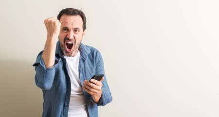 Senior man using smartphone annoyed and frustrated shouting with anger, crazy and yelling with...