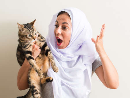 Arabian woman wearing white hijab holding a cat very happy and excited, winner expression celebrating victory screaming with big smile and raised handsの写真素材