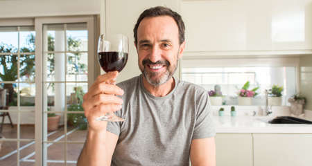 Middle age man drinking a glass of wine with a happy face standing and smiling with a confident...