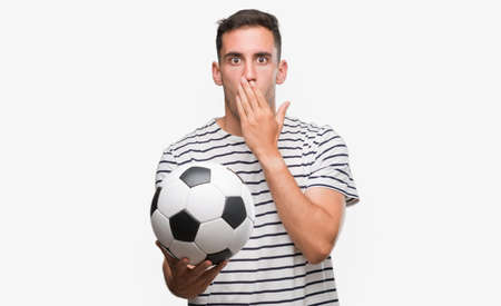 Handsome young man holding soccer football cover mouth with hand shocked with shame for mistake, expression of fear, scared in silence, secret conceptの写真素材