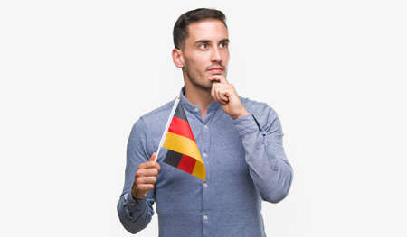 Handsome young man holding a flag of Germany serious face thinking about question, very confused...