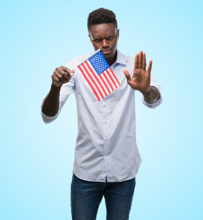 Young african american man holding USA flag with open hand doing stop sign with serious and confident expression, defense gestureの写真素材